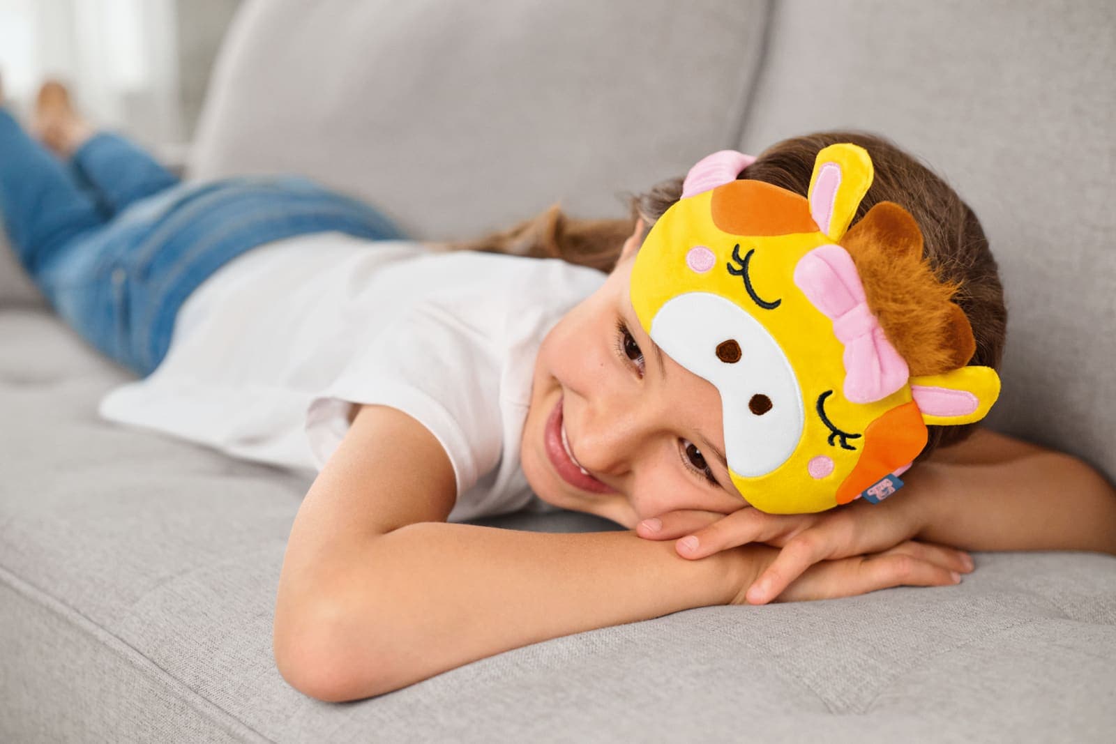 Girl wearing a yellow giraffe eye mask, smiling and lying on a sofa.