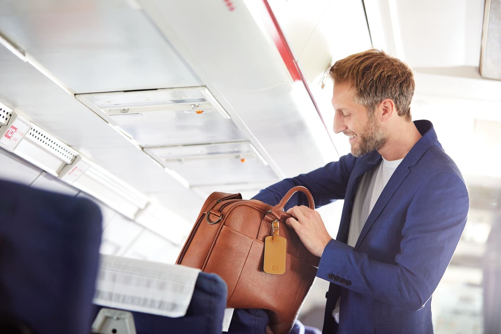 Man placing brown briefcase with luggage tag into airplane overhead compartment.