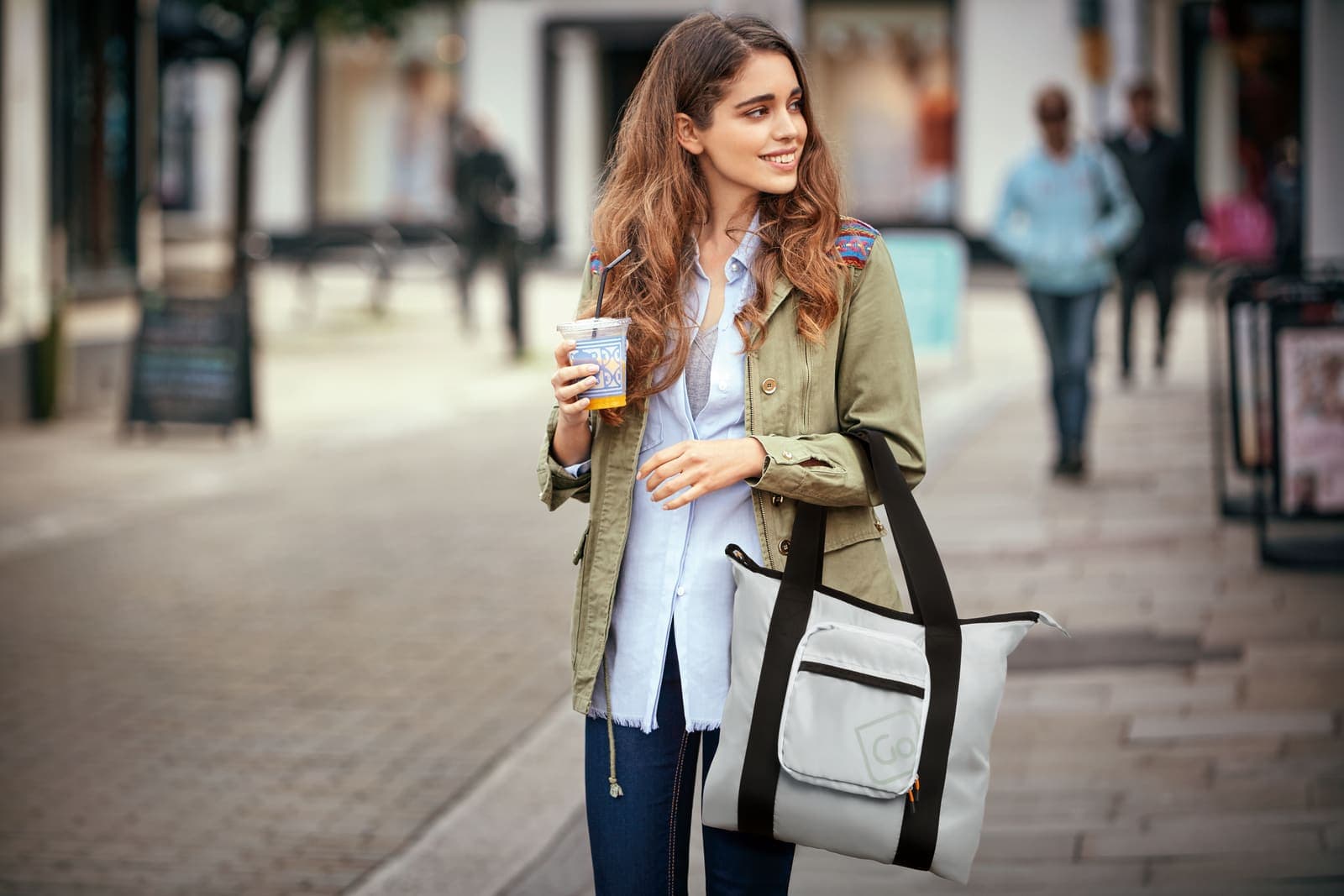 Woman holding a GO TRAVEL grey tote bag and a drink while walking on a city street.