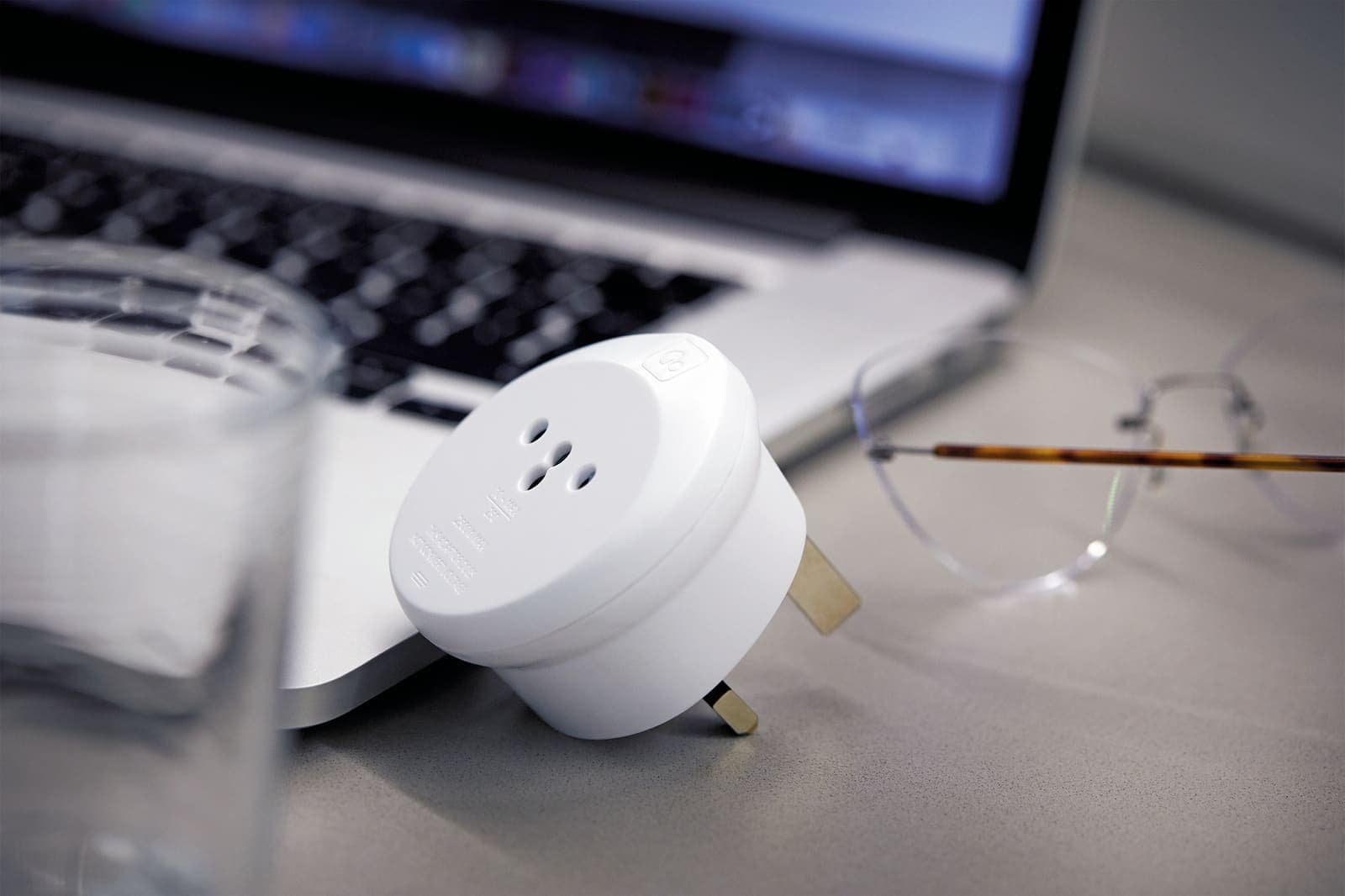 White GO TRAVEL Swiss/ITA-UK adaptor on a desk with a laptop and glasses.
