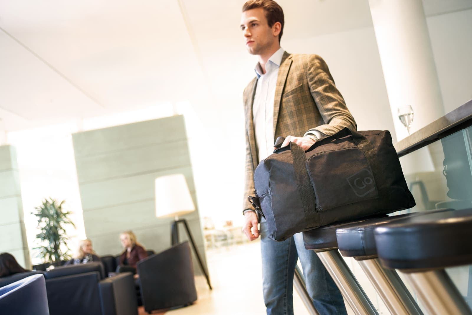 Man holding a black GO TRAVEL duffel bag in a modern lounge.