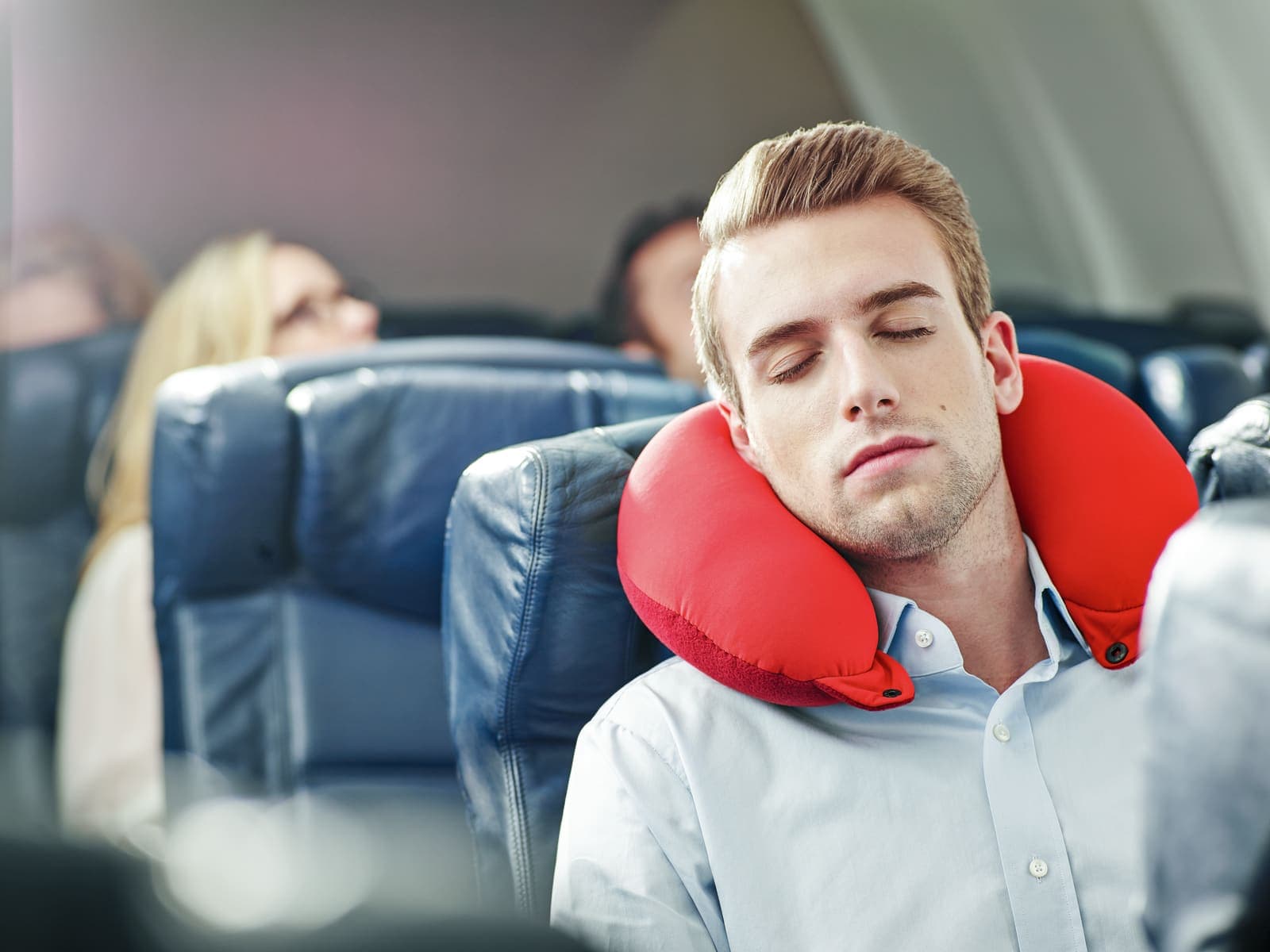 Man sleeping on airplane with red neck pillow.